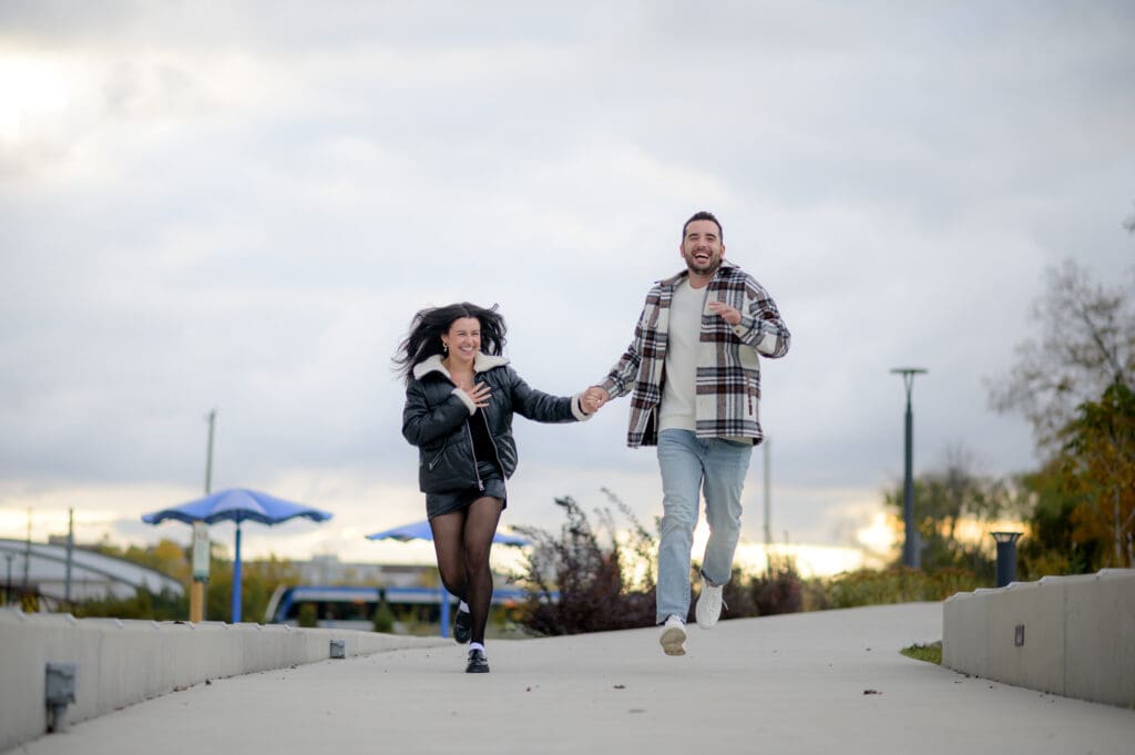 Engaged couple holding hands running in Waterloo Park with colorful fall trees in the background
