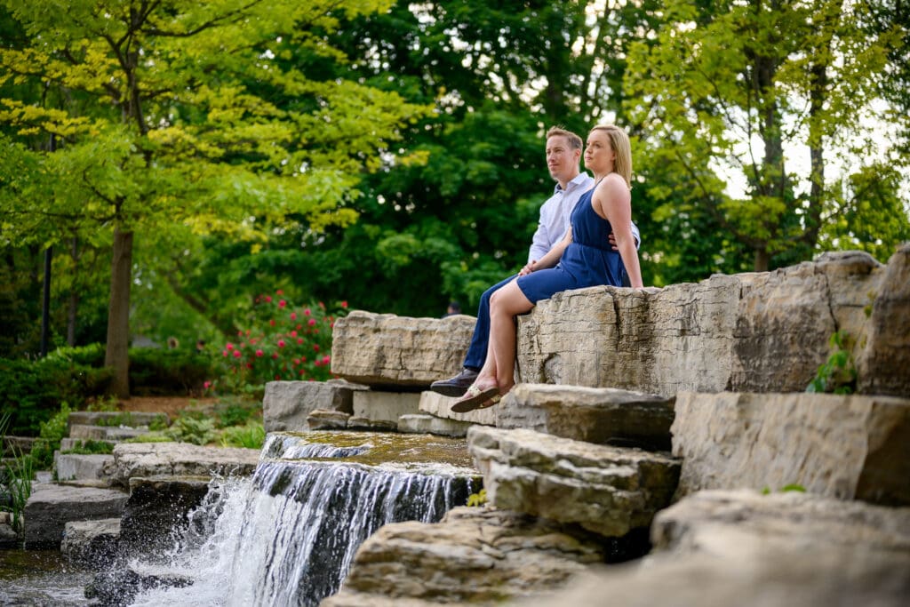 engaged couple holding hands sitting by waterfall at Victoria park Kitchener